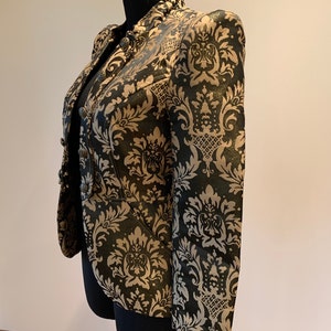 Pode incluir: Um blazer estruturado com um padr&atilde;o de damasco preto e dourado com acabamento cintilante. O casaco tem gola alta, mangas compridas e fecho frontal com bot&otilde;es met&aacute;licos redondos. O tecido apresenta um motivo floral ornamental repetido.