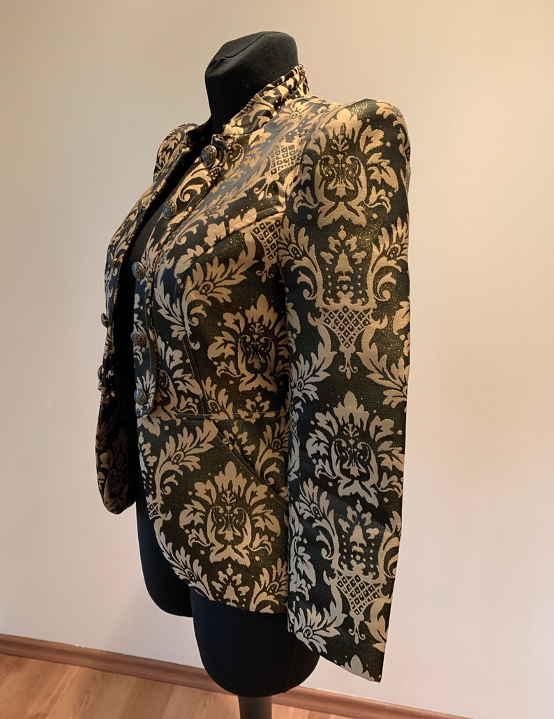 Pode incluir: Um blazer estruturado com um padr&atilde;o de damasco preto e dourado com acabamento cintilante. O casaco tem gola alta, mangas compridas e fecho frontal com bot&otilde;es met&aacute;licos redondos. O tecido apresenta um motivo floral ornamental repetido.
