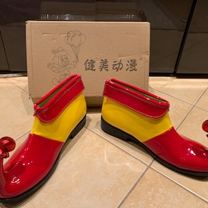 Könnte beinhalten: Ein Paar glänzende rot, gelb und grüne Clownschuhe mit gebogenen Zehen und einem roten Kugelornament. Die Schuhe stehen vor einem Pappkarton mit einer Cartoon-Clown-Illustration und chinesischem Text.