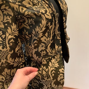 Pode incluir: Um blazer preto e dourado com um padr&atilde;o de damasco proeminente em todo o tecido. A pe&ccedil;a inclui uma gola estruturada, fecho frontal com bot&otilde;es met&aacute;licos ornamentados e uma abertura de bolso vis&iacute;vel na parte inferior frontal.