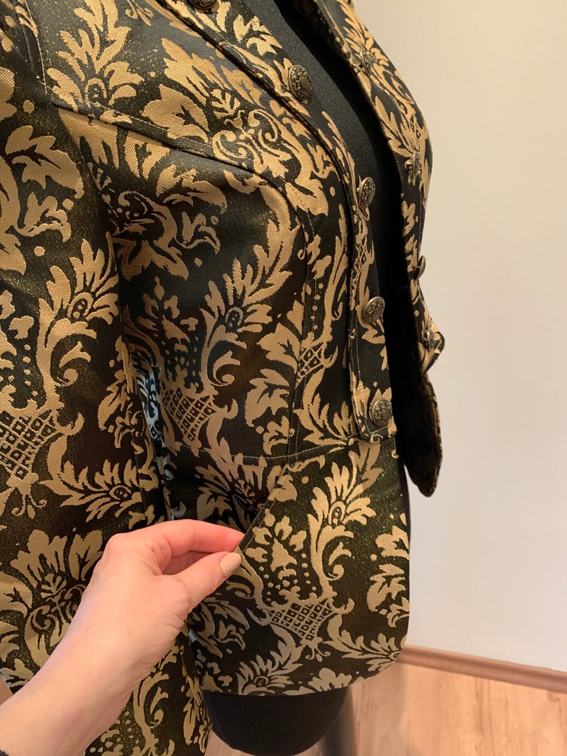 Pode incluir: Um blazer preto e dourado com um padr&atilde;o de damasco proeminente em todo o tecido. A pe&ccedil;a inclui uma gola estruturada, fecho frontal com bot&otilde;es met&aacute;licos ornamentados e uma abertura de bolso vis&iacute;vel na parte inferior frontal.