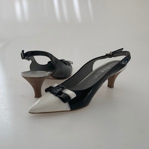 May include: Black and white slingback heels with a bow detail on the toe. The heels are made of patent leather and have a small block heel. The brand name "MARBE" is printed on the heel.
