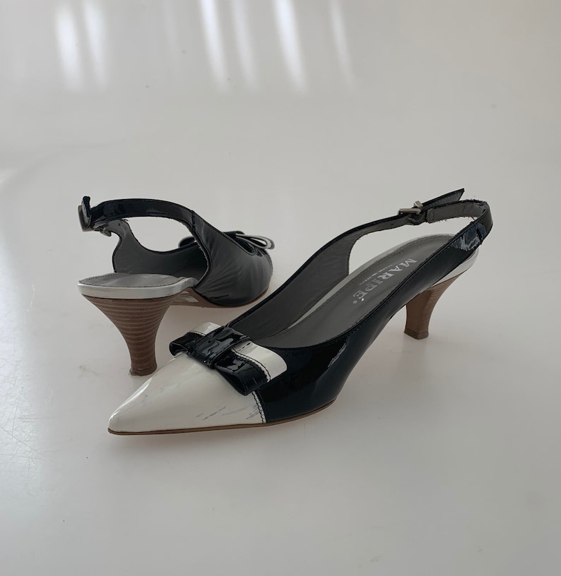 May include: Black and white slingback heels with a bow detail on the toe. The heels are made of patent leather and have a small block heel. The brand name "MARBE" is printed on the heel.