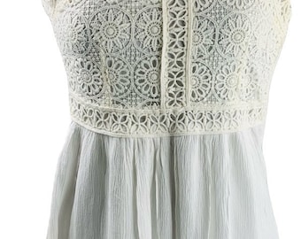Pode incluir: Uma blusa branca sem mangas com decote redondo e mangas curtas. A parte superior do corpete é feita de renda de crochê floral detalhada, enquanto a seção inferior consiste em um tecido leve e texturizado com uma bainha de renda a condizer.