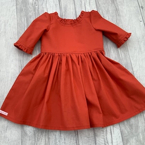 May include: A rust-coloured dress with a ruffled neckline and three-quarter sleeves. The dress has a gathered skirt and a small label on the side.
