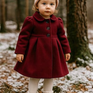 May include: A young child wearing a burgundy coat with a round collar and three black buttons. The coat has a flared skirt and is paired with a light pink beanie, white tights, and brown boots. The background shows a snowy forest.