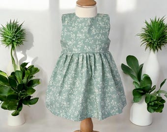 May include: A light green dress with a white floral pattern, displayed on a wooden mannequin. The dress has a sleeveless design and a gathered skirt. The background features white vases with green plants.