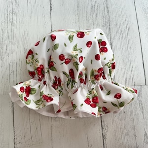 May include: A white fabric shower cap with a red cherry print. The cap has a ruffled edge and is gathered at the top.
