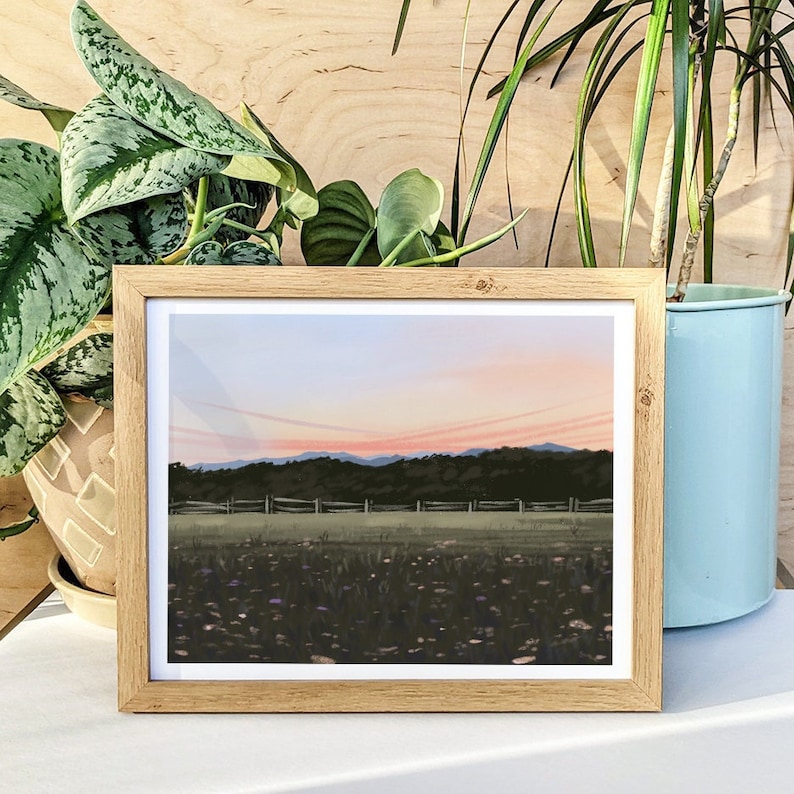 K&ouml;nnte beinhalten: Ein gerahmter Druck eines Landschaftsgem&auml;ldes, das eine Wiese mit Wildblumen im Vordergrund, einen Holzzaun und eine Reihe von B&auml;umen in der Ferne zeigt. Der Himmel ist in sanftem Rosa und Lila mit ein paar Wolken.