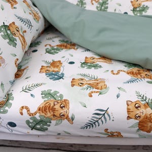 May include: Bedding set featuring a white base with a pattern of cartoon orange tabby cats, green leaves, and blue details. The top sheet is a solid sage green. The pillowcase and duvet cover are visible.