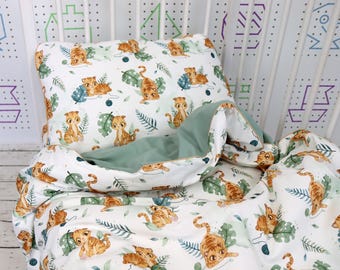May include: A white baby bedding set featuring a repeating pattern of orange tiger cubs and green leaves. The set includes a pillow and a blanket with a solid green interior. The bedding is placed inside a white crib.