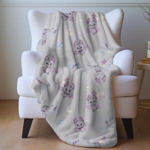 May include: A light gray blanket draped over a white armchair. The blanket features a repeating pattern of cartoon bats with purple wings, moons, stars, and floral accents. The blanket is made of a soft, plush material.