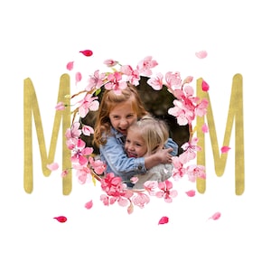 May include: Two young girls smiling and hugging, framed by a pink and white floral wreath with the word "MOM" in gold letters on either side.