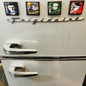 May include: A white refrigerator with silver handles and the word "Frigidaire" in cursive. Four rectangular magnets featuring classic horror movie posters of Dracula, The Wolf Man, Frankenstein, and The Bride of Frankenstein are attached to the refrigerator.