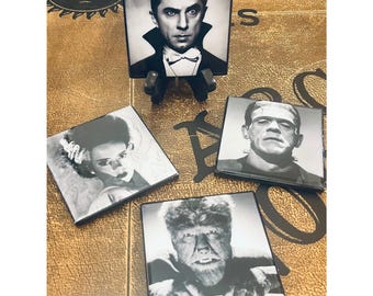 May include: A set of four square coasters featuring black and white portraits of classic horror movie characters including Dracula, Frankenstein's monster, the Bride of Frankenstein, and the Wolf Man, displayed on a textured brown surface.
