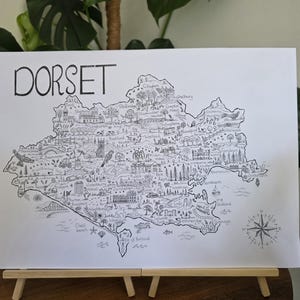 May include: A detailed black and white illustrated map of Dorset, England, featuring the word "DORSET" at the top. The map includes landmarks and towns, with a compass rose. The artwork is displayed on a small wooden easel.
