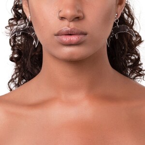 Może przedstawiać: Zbliżenie twarzy osoby z kolczykiem w nosie i dużymi srebrnymi kolczykami w kształcie skrzydeł. Osoba ma ciemnobrązowe włosy i nie ma makijażu.