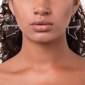 May include: A woman with curly brown hair wearing clear acrylic earrings shaped like a speech bubble with a ruler design.