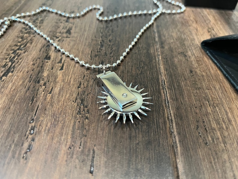 May include: A silver chain necklace with a pendant featuring a small, round, silver disc with a single diamond in the center. The disc is surrounded by a ring of sharp, pointed spikes.
