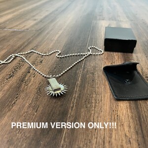 May include: A silver necklace with a circular pendant featuring a small, sharp blade. The necklace is displayed on a wooden surface with a black box and a black leather pouch.