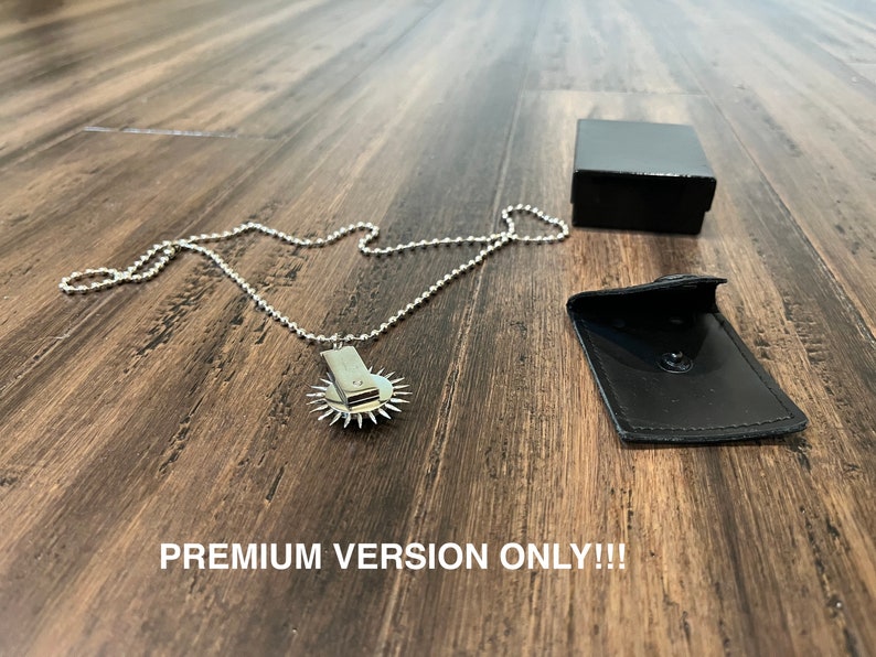 May include: A silver necklace with a circular pendant featuring a small, sharp blade. The necklace is displayed on a wooden surface with a black box and a black leather pouch.