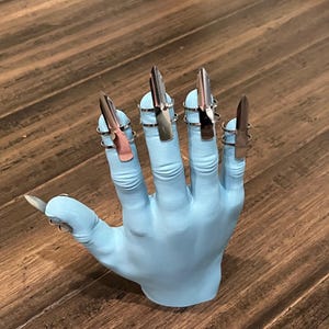 Peut inclure: Un modèle de main bleu clair avec cinq anneaux d'ongles en forme de griffes argentées.