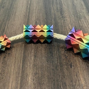 Puede incluir: Tres rodillos texturizados, con forma de púas, de color arcoíris están ensartados en una cuerda de color natural. Los rodillos son de metal y tienen un acabado arcoíris iridiscente.