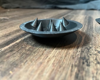 May include: A black, circular dish with six pointed spikes protruding from the center. The dish is made of a smooth, hard material and has a slightly textured surface.