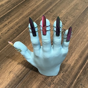May include: A white hand model with five iridescent, pointed, fake nails with gold rings on each finger.