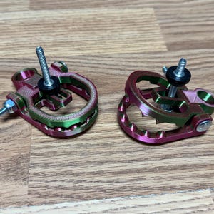 Op de afbeelding: Twee veelkleurige, 3D-geprinte componenten met schroeven en ringen. De componenten zijn voornamelijk roze en groen, met een cirkelvormig ontwerp en een centrale schroef. Ze liggen op een houten oppervlak.