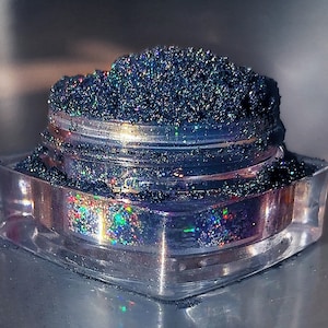 Pode incluir: Um pequeno frasco transparente cheio de glitter preto iridescente. O glitter está solto e tem um acabamento brilhante.