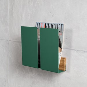 May include: A green metal magazine rack with two compartments. The rack is mounted to a wall and has a wooden base. A magazine titled "L'Officiel" is displayed in the rack.