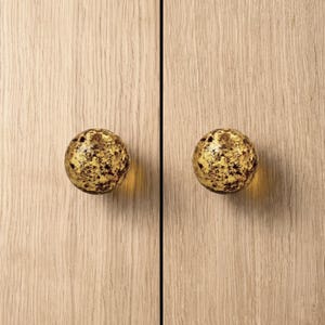 Pode incluir: Dois puxadores de armário esféricos com acabamento salpicado dourado e castanho. Os puxadores são montados numa porta de armário de madeira clara com grão vertical. Um elemento decorativo.