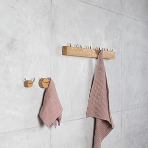 May include: A wooden wall-mounted coat rack with five gold hooks and two smaller wooden hooks with silver rings. A pink towel hangs on one of the gold hooks.