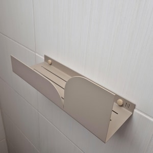 May include: A beige metal shelf with rounded edges and a small, rectangular cutout. The shelf is mounted to a white tiled wall with wooden screws. The shelf has a small, engraved logo that says "N".