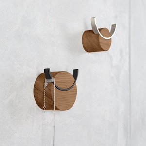 Op de afbeelding: Twee houten wandhaken met metalen details. Eén haak heeft een zwarte metalen haak, de andere een zilveren metalen haak. Een zilveren ketting hangt aan de zwarte haak. De haken zijn bevestigd aan een lichtgrijze muur.