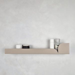 Puede incluir: Una estantería de metal marrón claro con bordes redondeados, con una botella blanca, un tarro negro y un tarro blanco en la parte superior. La estantería está montada en una pared blanca.