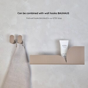 K&ouml;nnte beinhalten: Ein beiges Wandregal und zwei passende ovale Haken. Eine wei&szlig;e Tube ESPA Body Wash steht auf dem Regal. Ein beiges Handtuch h&auml;ngt an den Haken. Der Text auf dem Bild lautet "Can be combined with wall hooks BAUHAUS".