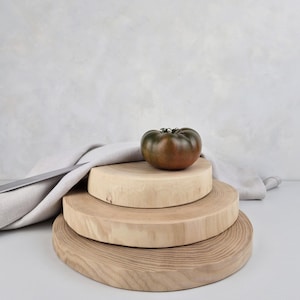 May include: Three round wooden chopping boards stacked on top of each other. The top board has a dark green tomato on it. The boards are made of light wood with a natural grain.