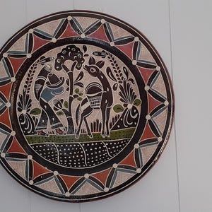 May include: Round decorative plate with a detailed illustration. The plate features a central scene with figures and animals, surrounded by a geometric border in red, black, and cream colors. The plate is mounted on a white wall.