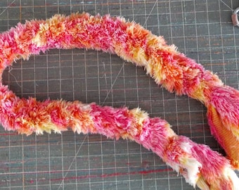 May include: A vibrant, handmade lei in shades of pink, orange, and yellow. The fluffy, textured garland is arranged in a circular shape, with a tail-like end. Scissors are visible in the background.