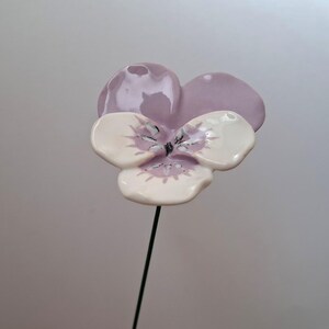 Pode incluir: Uma flor de amor-perfeito de cer&acirc;mica com a parte superior lil&aacute;s claro e p&eacute;talas brancas com detalhes em roxo. A flor &eacute; presa a uma haste fina de arame verde escuro, ideal para decora&ccedil;&atilde;o de jardim ou arranjos florais.