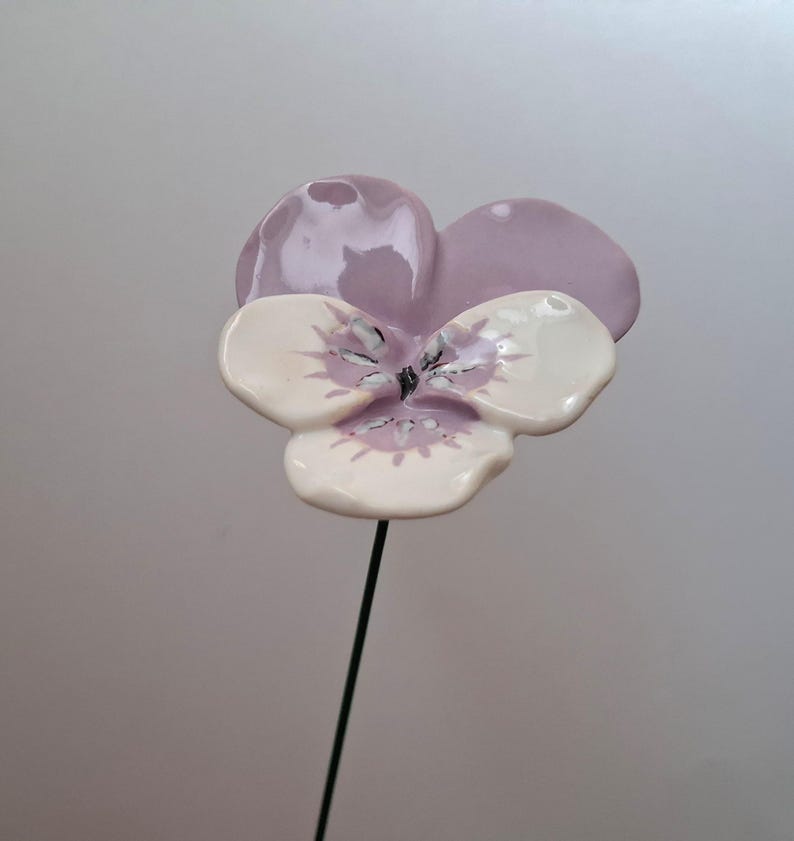 Pode incluir: Uma flor de amor-perfeito de cer&acirc;mica com a parte superior lil&aacute;s claro e p&eacute;talas brancas com detalhes em roxo. A flor &eacute; presa a uma haste fina de arame verde escuro, ideal para decora&ccedil;&atilde;o de jardim ou arranjos florais.