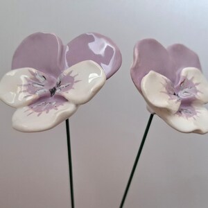 Pode incluir: Duas flores de amor-perfeito de cer&acirc;mica com p&eacute;talas de lavanda e centros brancos e lavanda. As flores s&atilde;o montadas em hastes de arame verde, perfeitas para arranjos florais ou artesanato. Os amores-perfeitos t&ecirc;m um acabamento brilhante.