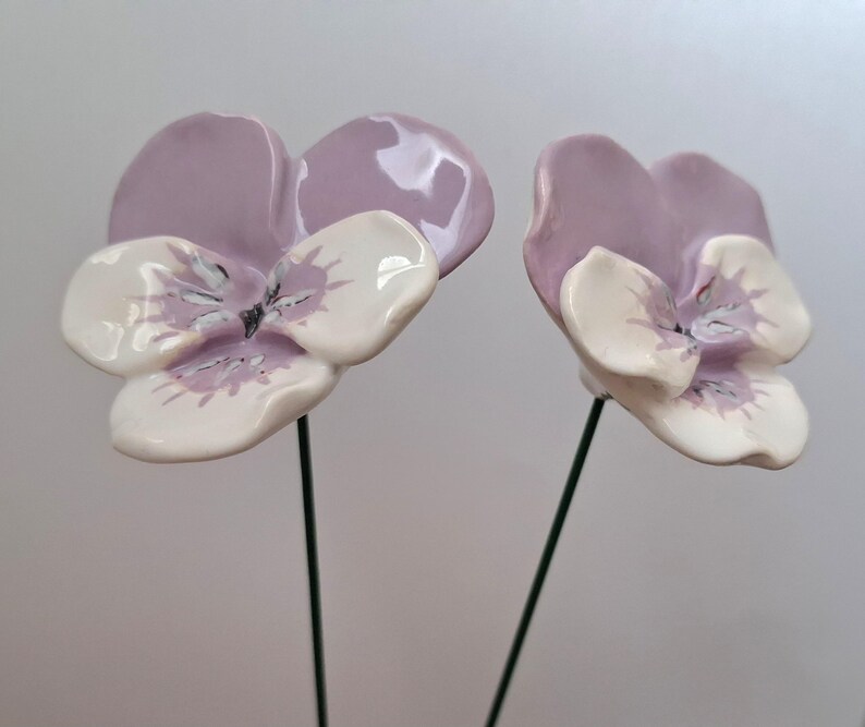 Pode incluir: Duas flores de amor-perfeito de cer&acirc;mica com p&eacute;talas de lavanda e centros brancos e lavanda. As flores s&atilde;o montadas em hastes de arame verde, perfeitas para arranjos florais ou artesanato. Os amores-perfeitos t&ecirc;m um acabamento brilhante.