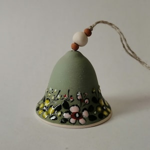May include: A green ceramic bell with a floral design in yellow, pink, black, and white. The bell has a brown and white bead string attached to the top.