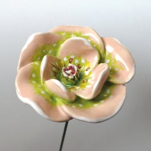 May include: A ceramic flower sculpture with a light pink and green color scheme. The flower has multiple petals with white polka dots and a dark red center. The sculpture is mounted on a thin, black metal stem.