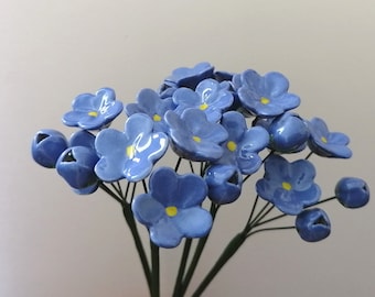 May include: A bouquet of ceramic forget-me-nots. The flowers are a light blue with yellow centers, and the buds are a darker blue. The stems are thin and dark green. The flowers are arranged in a bunch.