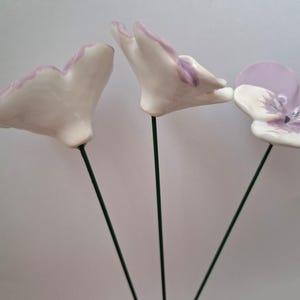 Pode incluir: Tr&ecirc;s esculturas de flores de cer&acirc;mica com p&eacute;talas brancas com bordas lavanda, montadas em hastes de arame verde. Uma flor est&aacute; totalmente aberta, exibindo um centro lavanda e branco. As outras duas est&atilde;o em processo de abertura.