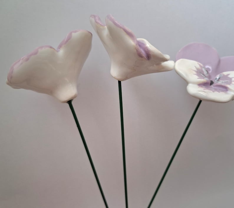 Pode incluir: Tr&ecirc;s esculturas de flores de cer&acirc;mica com p&eacute;talas brancas com bordas lavanda, montadas em hastes de arame verde. Uma flor est&aacute; totalmente aberta, exibindo um centro lavanda e branco. As outras duas est&atilde;o em processo de abertura.